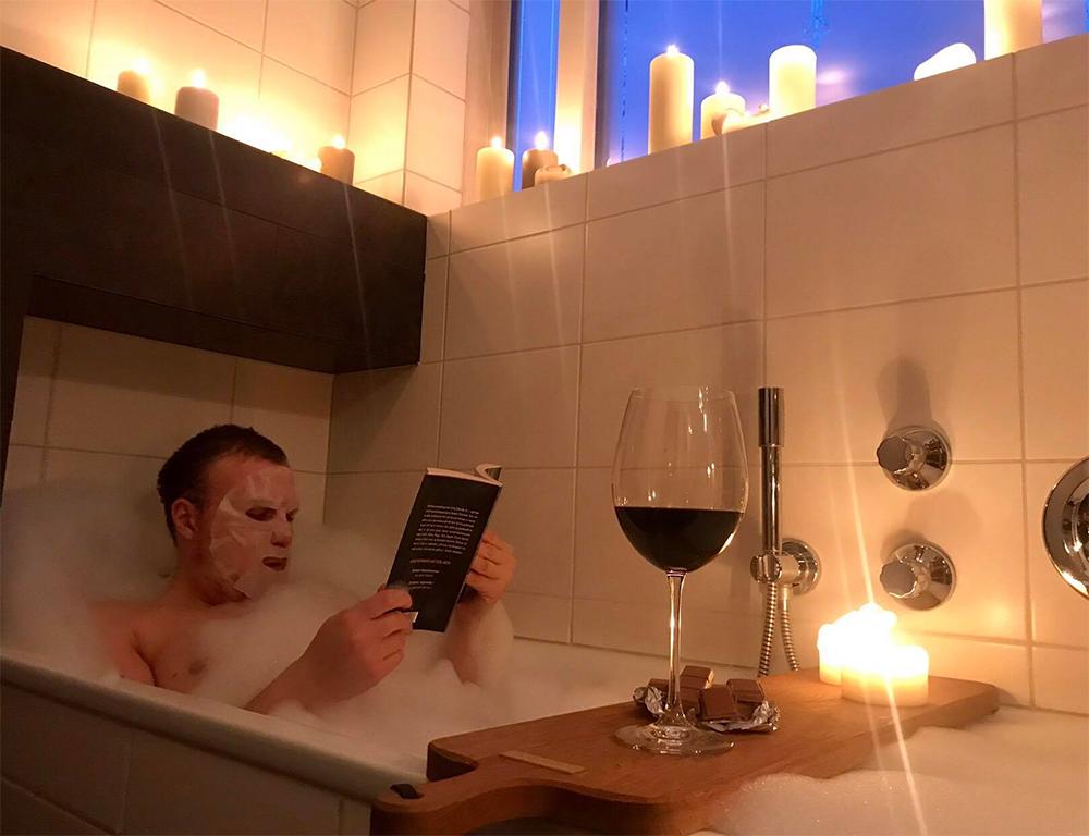 Mynd af manni í baði við kertaljós, með andlitsmaska og rauðvín í glasi.
