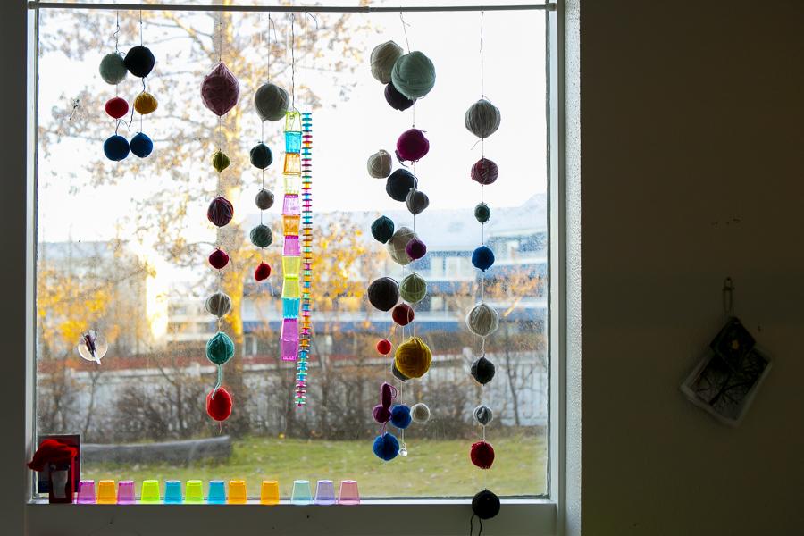 Hangandi litríkt skraut úr garnhnyklum og plastglösum í glugga.