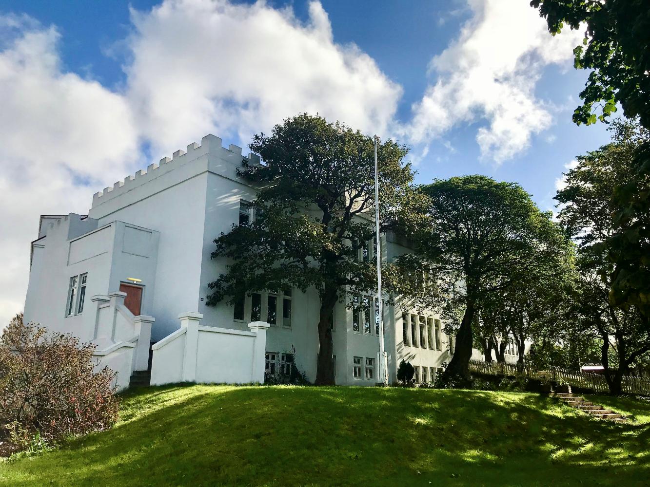 Hús Söngskólans í Reykjavík séð utanfrá. Stór tré í garðinum, hvítt hús og blár himinn.