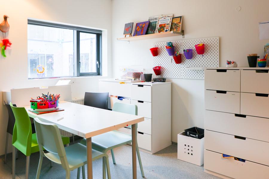 Room with window, table, chairs and storage cabinets. Boxes of paints sit on the table while various craft supplies are stored on the cabinets and wall. 