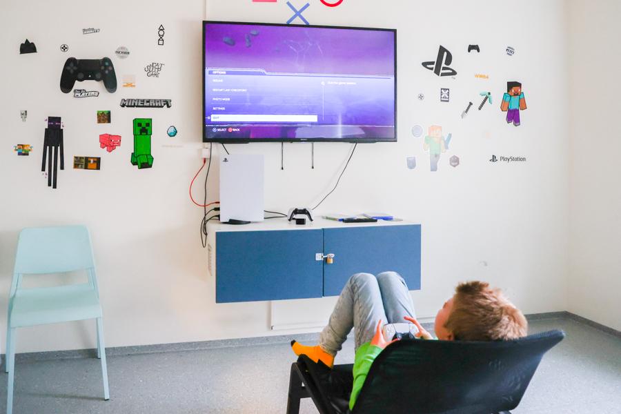 Child sits in a chair looking at the wall-mounted screen. Video game-related pictures hang on the wall. Cabinets are positioned below the screen. 