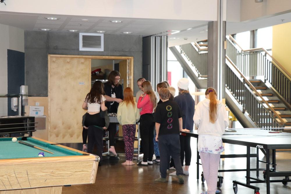 Children gathered in a group talking with a staff member. Stairs to the side. A pool table and ping pong table in the foreground. 
